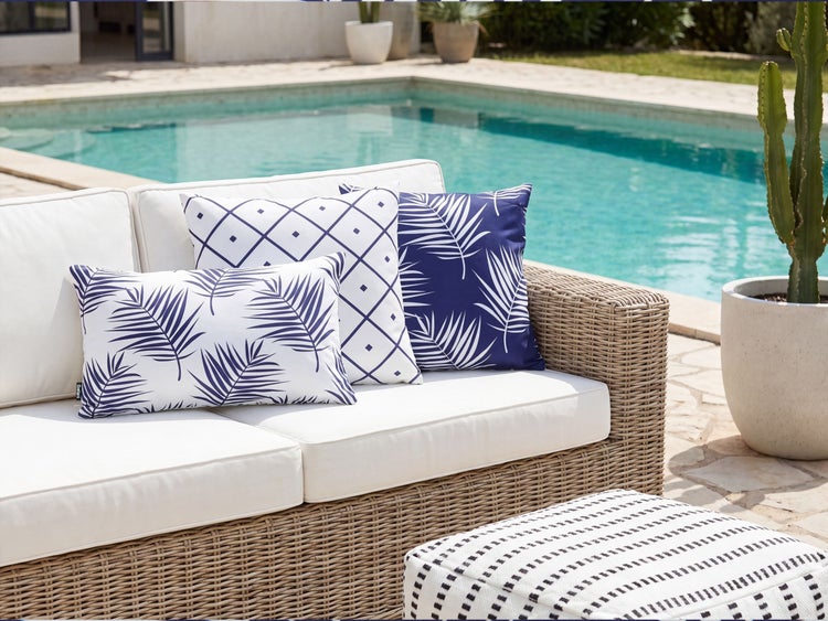 Gartenlounge aus Rattan mit weissen Sitzpolstern und verschiedenen Zierkissen vor einem Swimmingpool.