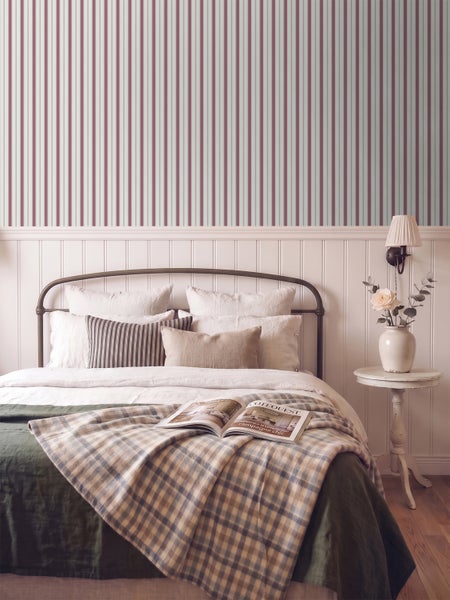 Helles Schlafzimmer im Landhausstil mit gestreifter Tapete, weißer Holzvertäfelung, Metallbett und einem Beistelltisch.