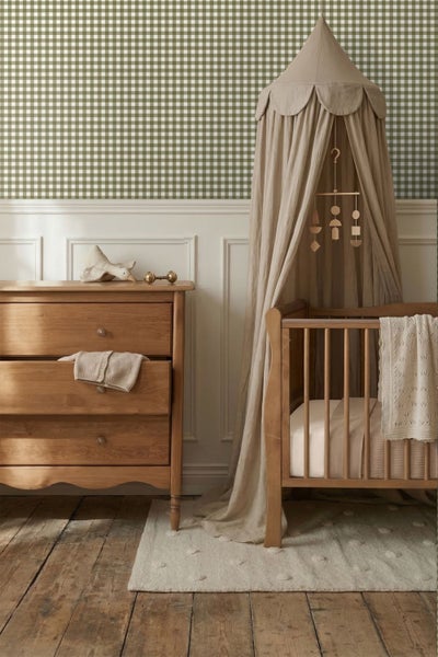Babyzimmer mit Holzkommode, Gitterbett mit beigem Betthimmel und grün-weiß karierter Tapete über weißer Wandvertäfelung.