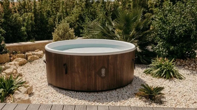 Runder Whirlpool mit Außenwand in Holzoptik, aufgestellt in einem Garten auf einer Kiesfläche, umgeben von mediterranen Pflanzen.