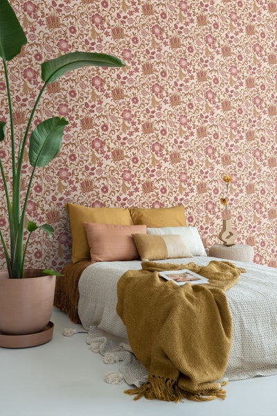 Helles Schlafzimmer mit Blumentapete, Bett mit strukturierten Kissen, gelber Strickdecke und einer großen Zimmerpflanze im Topf.