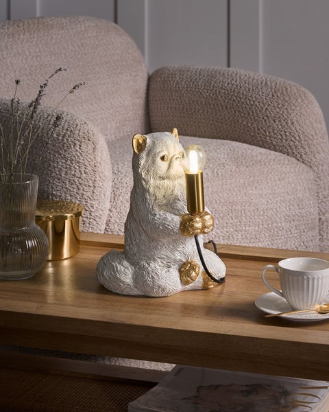 Dekorative Tischleuchte als weiße Katze mit Golddetails auf einem Holztisch neben einer Tasse und Vase mit Trockenblumen vor hellem Sessel.