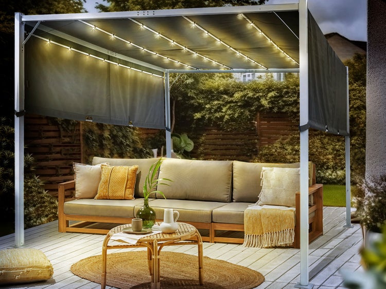 Pergola mit LED-Beleuchtung über einer Lounge-Sitzgruppe aus Holz auf einer Terrasse im Garten bei Dämmerung.