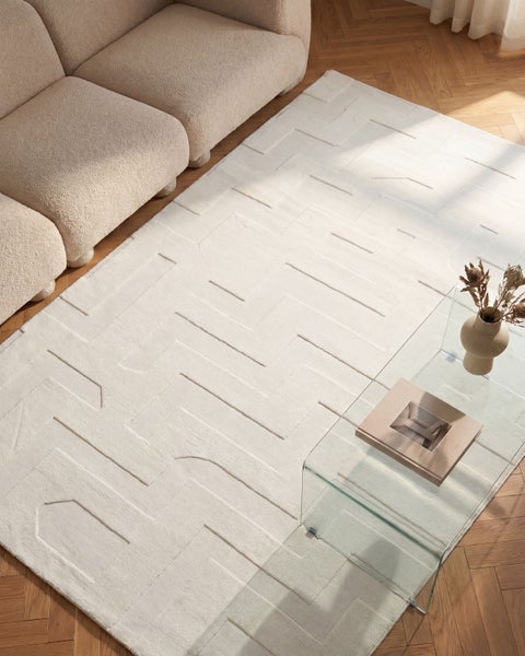 Wohnzimmer mit einem großen Teppich mit geometrischem Reliefmuster auf Parkettboden neben einem Sofa aus Boucléstoff und einem Glastisch.