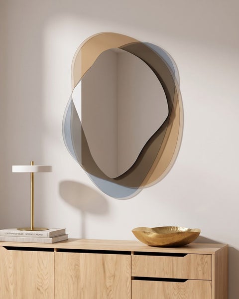 Wandspiegel mit asymmetrischem, mehrlagigem Design in Glasoptik über einer Holzkommode mit Tischlampe und Deko-Schale.