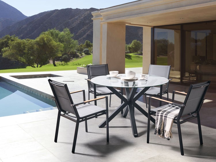 Terrasse mit runder Glasplatte, vier Stühlen, Schwimmbad und Berglandschaft im Hintergrund.