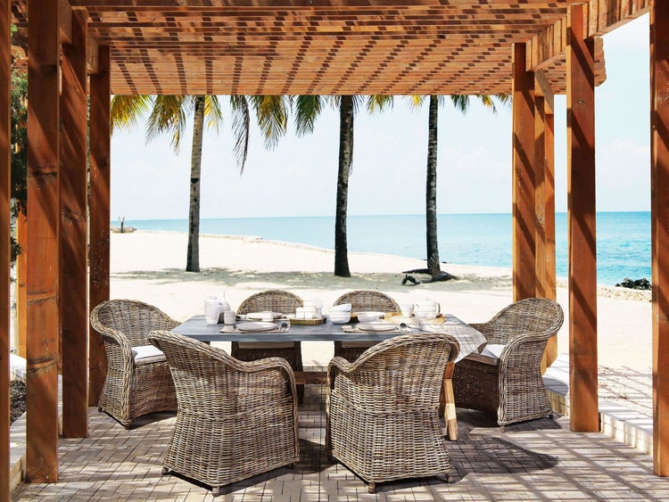 Gedeckter Tisch und Rattanmöbel unter einer Holzpergola mit Blick auf den Strand.