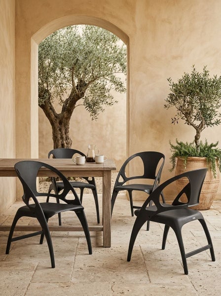 Moderne Gartenmöbelgruppe mit Esstisch aus Holz und vier Stühlen aus Kunststoff auf einer Terrasse mit Olivenbäumen und mediterranem Ambiente.