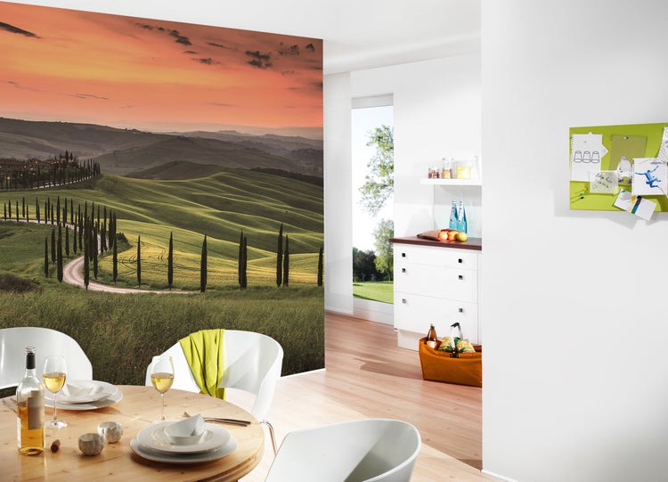 Fototapete mit Toskana-Landschaft bei Sonnenuntergang in einem hellen, modern eingerichteten Esszimmer mit Holztisch und weißen Stühlen.