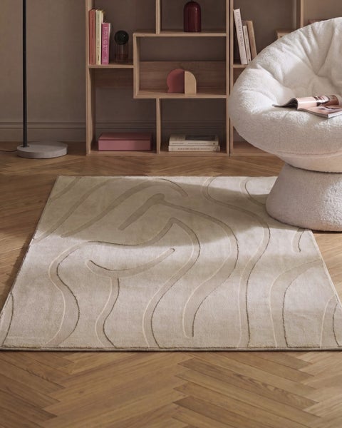 Rechteckiger Teppich mit modernem Reliefmuster in einem Wohnzimmer mit Holzregal und gemütlichem Sessel auf einem Parkettboden.