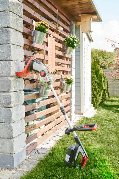 Akku Rasentrimmer GT 4030 stehend vor einer Holzwand mit Blumenkübeln in einem Garten.