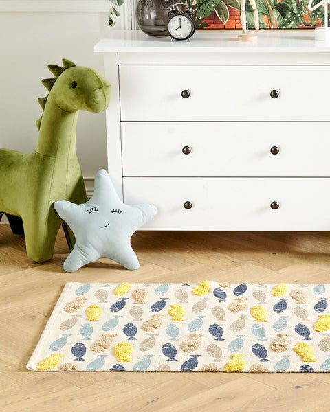 Rechteckiger Kinderteppich mit bunten Fischmotiven vor einer weißen Kommode, einem grünen Plüsch-Dinosaurier und einem hellblauen Sternkissen.