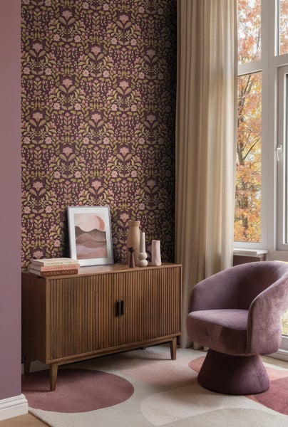 Vliestapete mit symmetrischem Blumenmuster im Vintage-Stil in einem Wohnzimmer mit Holzkommode, lila Sessel und gemustertem Teppich.