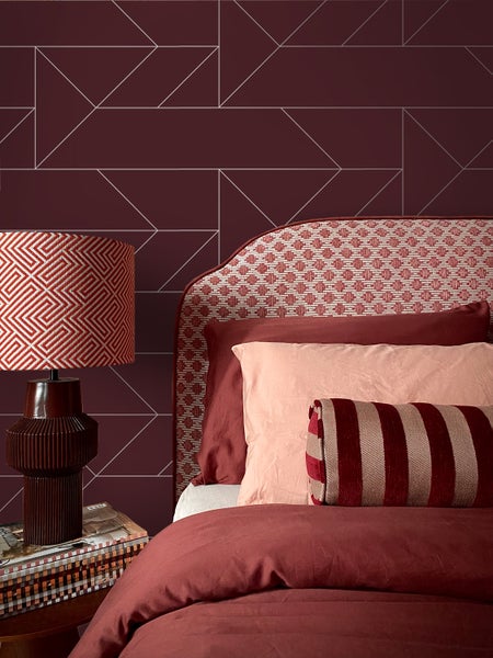 Vlies-Tapete mit geometrischem Muster in Dunkelrot und Silber, abgebildet in einem Schlafzimmer mit passender Bettwäsche und Tischlampe.