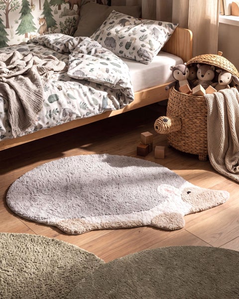 Kinderzimmer mit einem Igel Teppich auf Holzboden, neben einem Bett und einem geflochtenen Korb mit Stofftieren und Holzbausteinen.