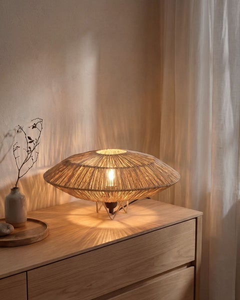 Tischleuchte mit flachem, rundem Schirm aus Kordelgeflecht auf einer Holzkommode. Das Licht wirft ein radiales Muster an die Wand.