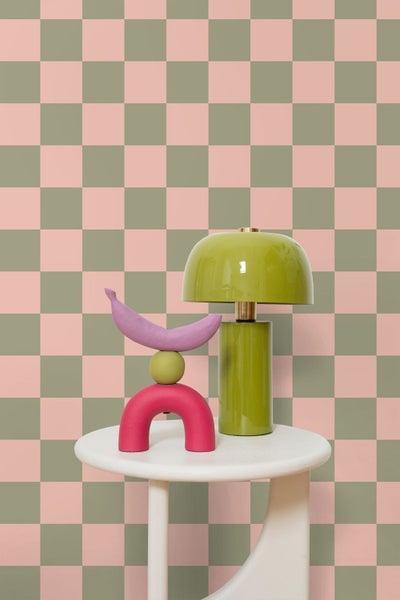 Vliestapete mit Karomuster in Olivgrün und Rosa hinter einem weißen Beistelltisch mit grüner Tischleuchte und bunter Skulptur.