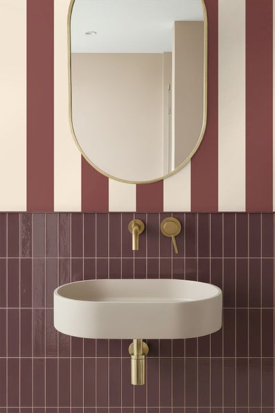 Modernes Badezimmer mit ovalem Waschbecken, goldfarbener Wandarmatur, kapselförmigem Spiegel, gestreifter Tapete und vertikalen dunkelroten Fliesen.