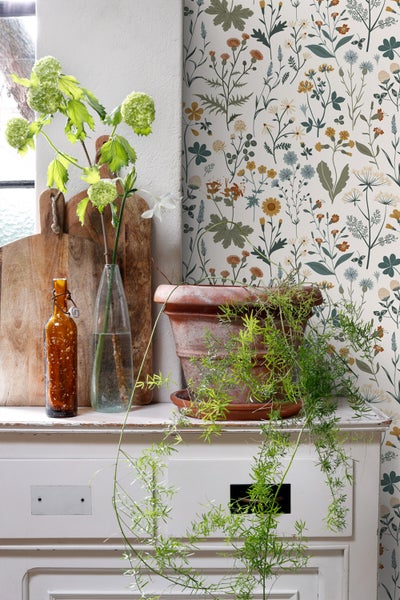 Dekorative Szene mit Blumentapete, Topfpflanze, Vasen und Holzbrettern auf einer weißen Kommode