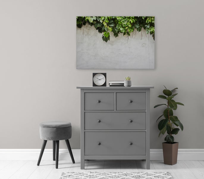 Eine Kommode mit vier Schubladen steht vor einer Wand mit einem Wandbild, daneben ein Hocker, eine Pflanze und ein kleiner Teppich.