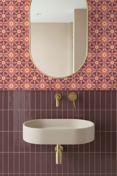 Modern gestaltetes Badezimmer mit gemusterter Tapete, dunklen vertikalen Fliesen, ovalem Spiegel und beigefarbenem Waschbecken mit Goldarmaturen.