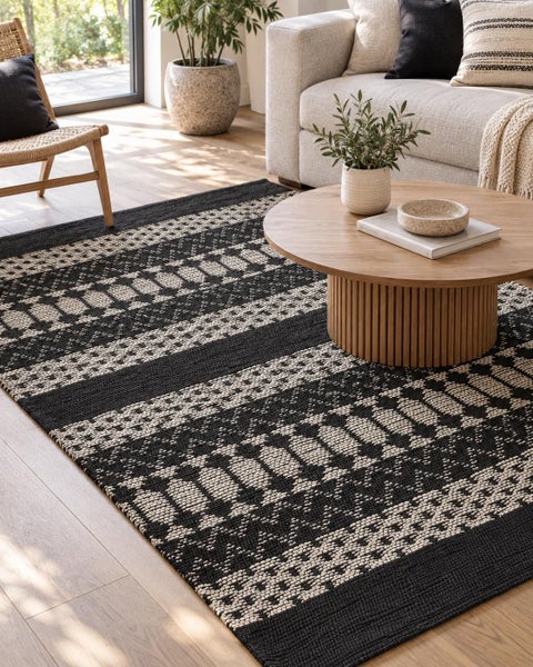 Gewebter Teppich mit geometrischem Muster in einem hellen Wohnzimmer mit Sofa und Couchtisch aus Holz.