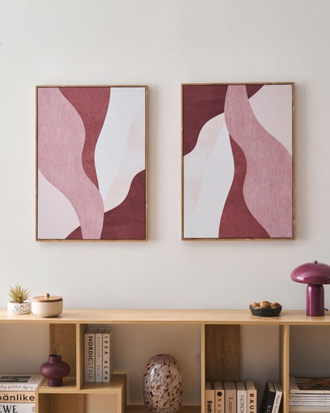 Zwei abstrakte Leinwandbilder mit wellenförmigen Mustern in Rosa und Burgunderrot mit Holzrahmen über einem Holzregal.