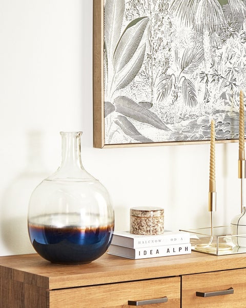 Dekoratives Arrangement mit einer Glasvase, Kerzen, Büchern und einem gerahmten botanischen Druck auf einem Holzschrank