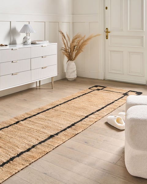 Langer Jute-Läufer mit schwarzen geometrischen Bordüren auf hellem Holzboden in einem hellen Zimmer neben einer weißen Kommode.