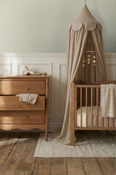 Hell eingerichtetes Kinderzimmer mit karierter Vliestapete, Holzkommode und Babybett mit Stoffhimmel auf einem Dielenboden.