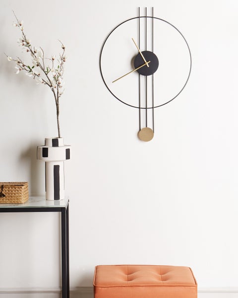 Moderne minimalistische Wanduhr mit Metallrahmen, Goldzeigern und goldenem Pendel neben einer Vase mit Zweigen und einem Beistelltisch.