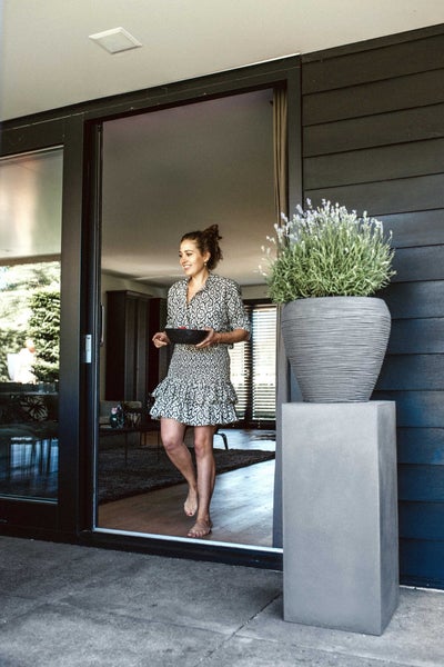 Moderne Terrasse mit Frau an einer Schiebetür, ein strukturierter Pflanzkübel mit Lavendel auf einem hohen Sockel in Betonoptik.