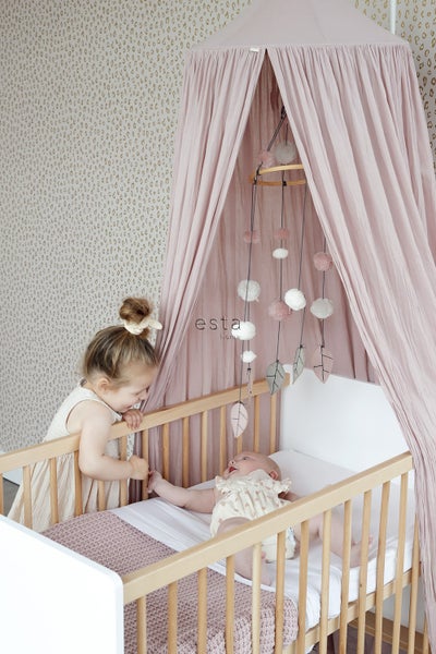 Szene mit einem Babybett mit Himmel und einem Mobile mit Blättern und Pompons. Ein Kind steht am Bett.