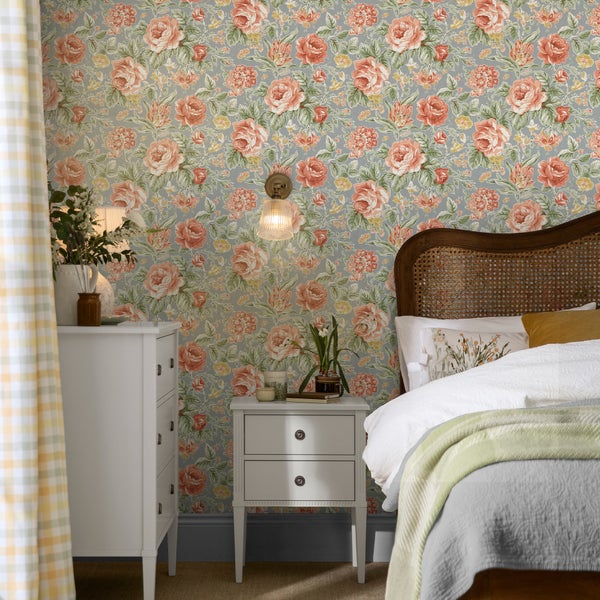 Schlafzimmer mit blumiger Tapete mit Rosenmuster, weißer Kommode, Nachttisch, Bett mit Rattan Kopfteil und Wandlampe in rustikalem Landhausstil.