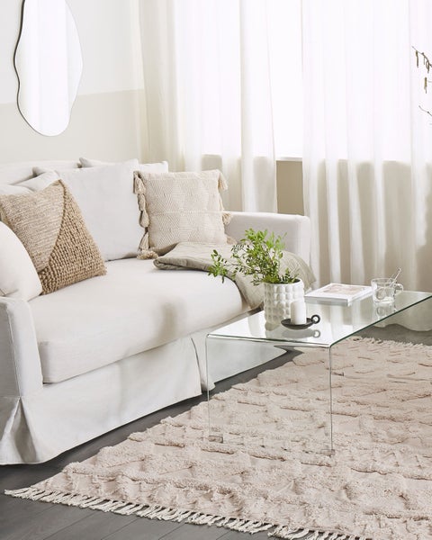 Helles Wohnzimmer mit hellem Sofa, Zierkissen, einem Glastisch und einem strukturierten Teppich mit Rautenmuster und Fransen.