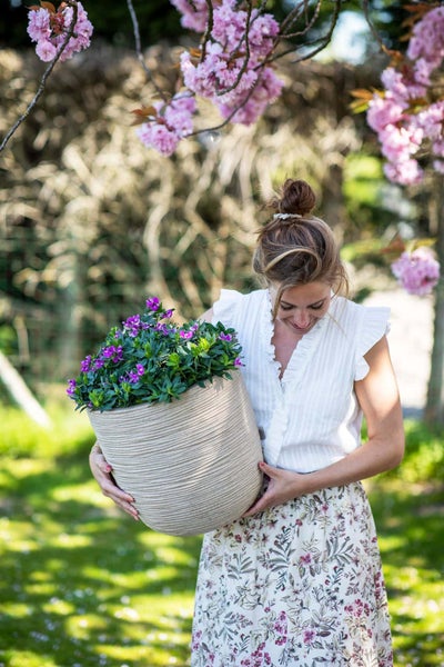 Eine Frau hält einen großen, runden Pflanztopf mit horizontalen Rillen und violetten Blumen in einem sonnigen Garten.