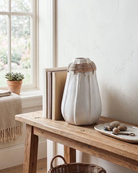 Dekorative Vase aus Keramik mit Rattanumwicklung, stehend auf einem Holztisch neben Büchern und einer Ablageschale mit Steinen vor einem Fenster.