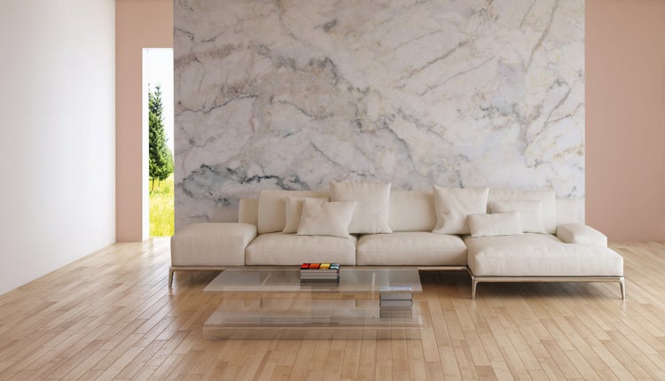 Modernes Wohnzimmer mit heller Eckcouch, Glastisch und einer großen Wand in Marmoroptik über hellem Holzfußboden.