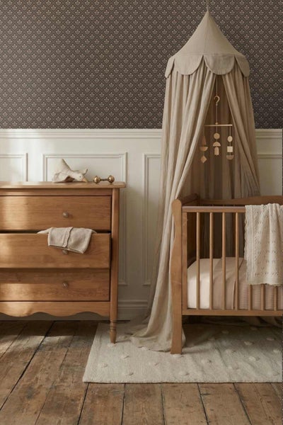 Kinderzimmer mit dunkler gemusterter Tapete, weißer Wandtäfelung, Holzkommode und Babybett mit Stoffhimmel über einem Holzboden.