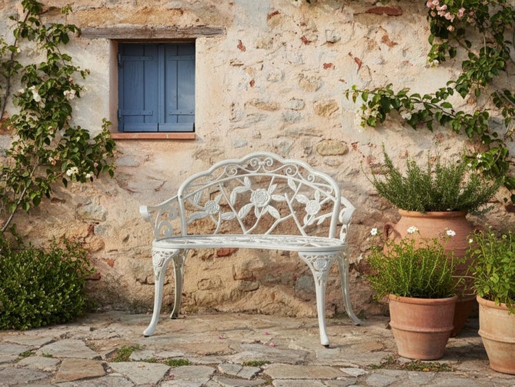 Eine Gartenbank aus Metall steht vor einer Steinwand mit Fenster und Pflanzen.