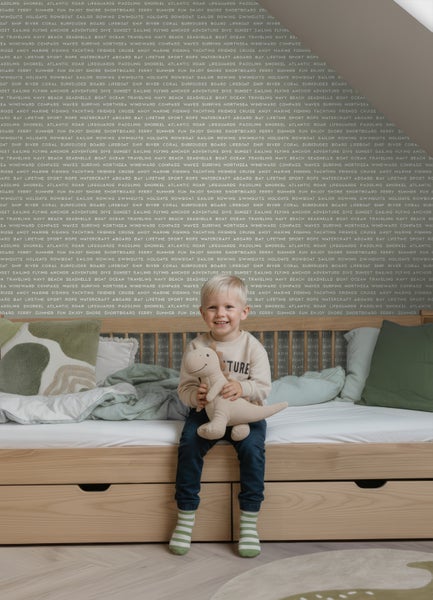 Hell gestaltetes Kinderzimmer mit einem Funktionsbett aus Holz mit Schubladen, einem Kind mit Plüschtier und einer Tapete mit Textmuster.