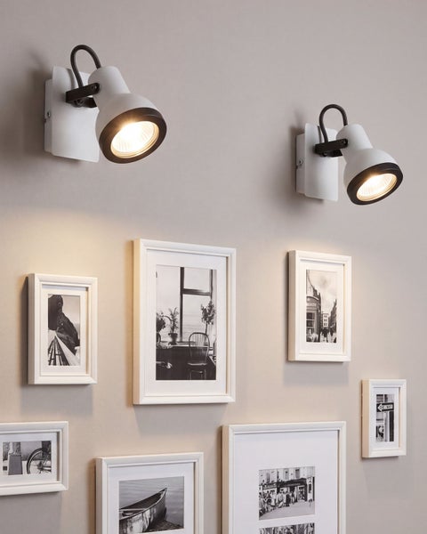Zwei weiße Wandstrahler beleuchten eine Galerie aus verschiedenen schwarz-weißen Fotografien in weißen Rahmen an einer hellen Wand.