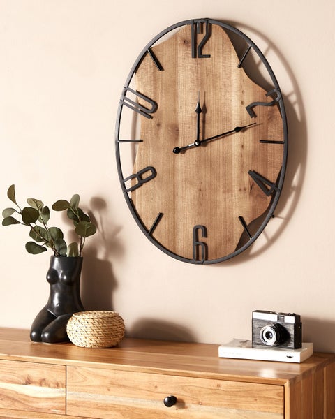 Ovale Wanduhr aus Naturholz und schwarzem Metall in modernem Design, aufgehängt in einem hellen, organisierten Raum über einer Kommode aus Holz.