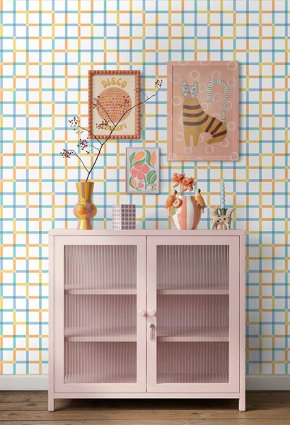 Hell gestalteter Raum mit einem rosa Metallschrank mit Glastüren vor einer bunt karierten Tapete sowie diversen Kunstwerken und Vasen.