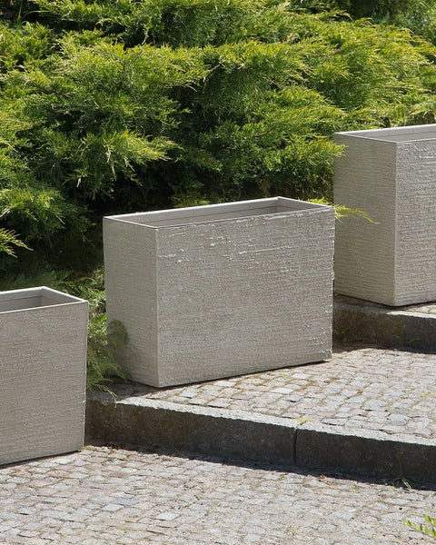 Rechteckige Pflanzgefäße aus Beton auf Steinstufen vor grünen Büschen.