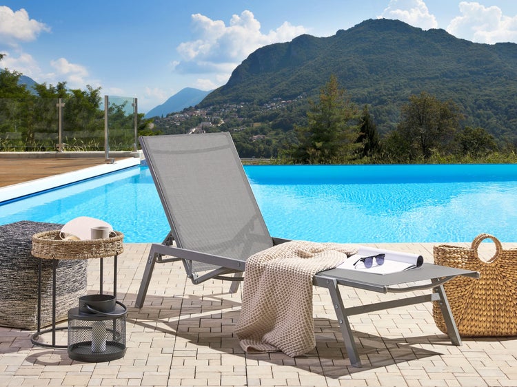 Szene mit Sonnenliege, Beistelltisch, Pouf, Laterne, Strandtasche und Pool vor Bergkulisse.