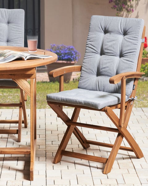 Klappbarer Gartenstuhl aus Holz mit grauer Hochlehner-Auflage und Armlehnen neben einem runden Holztisch auf einer gepflasterten Terrasse.