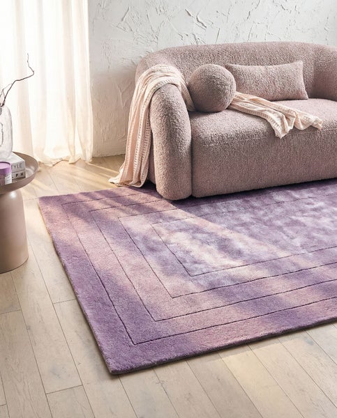 Modernes helles Wohnzimmer mit einem rechteckigen lilafarbenen Teppich mit geometrischem Linienmuster auf Holzboden neben einem Polstersofa.