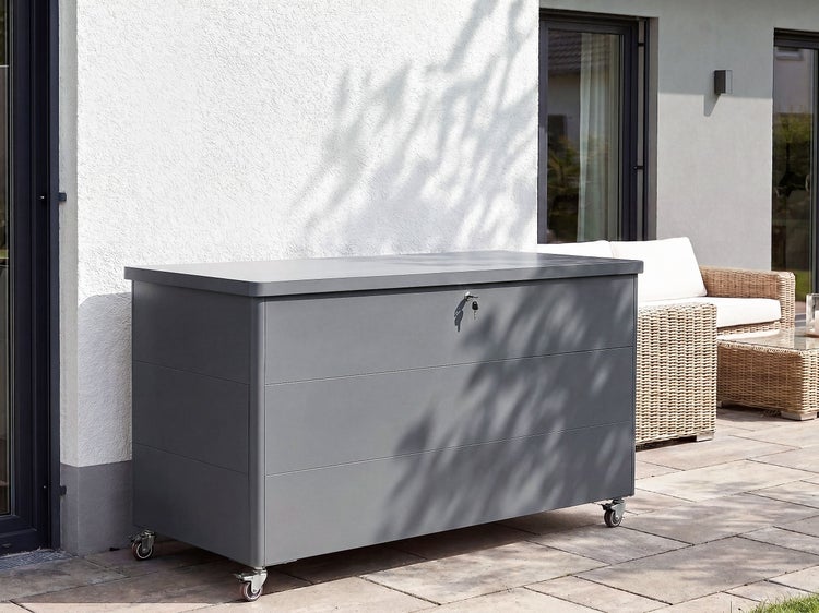 Eine abschließbare Kissentruhe aus Metall auf Rollen steht auf einer Terrasse vor einer Hauswand.