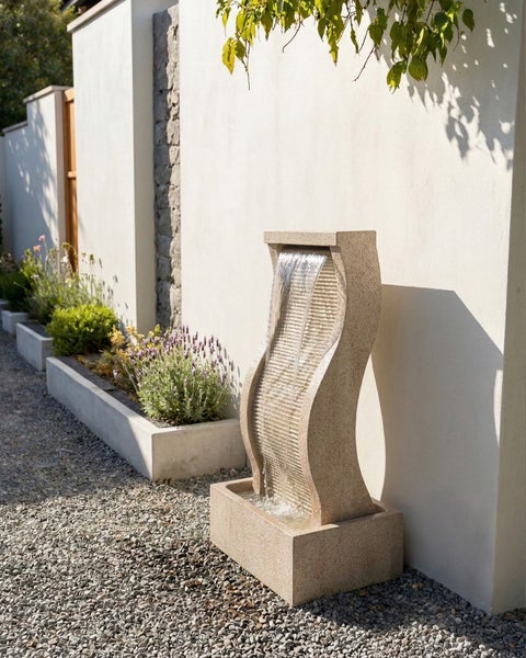 Moderner Gartenbrunnen aus Stein in Wellenform an einer weißen Wand mit Kiesuntergrund und Pflanzen.
